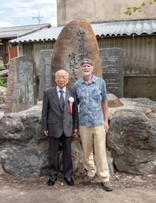 Hiroshi Doi Sensei and William Lee Rand stand together in front of large engraved stone monuments outdoors, one in a suit with a ribbon and the other in casual clothes and a cap.