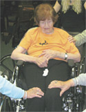 An elderly woman in a wheelchair receives supportive hand-on-shoulder contact from others in a group setting.