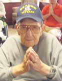 An elderly veteran in a baseball cap sits with his hands pressed together in a prayer-like meditative pose.