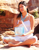 A woman in white sitting outdoors, playing a translucent blue crystal singing bowl for sound healing.