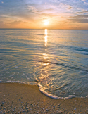 A peaceful ocean sunrise with gentle waves washing onto a sandy shore under a cloudy golden sky.