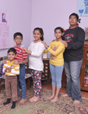 Five children of various ages standing in a line on a patterned rug, smiling and posing for a group photo indoors.