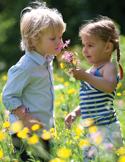 A young boy and girl standing in a field of yellow wildflowers, gently leaning in to smell a pink flower.