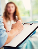 Close-up of a person’s hand writing on a clipboard with a woman sitting in the blurred background.