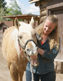 A smiling woman gently pets the face of a light-colored horse showing its teeth in a rustic outdoor setting.