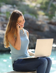 A smiling woman sits outdoors by the water, talking on a cell phone while working on a laptop.