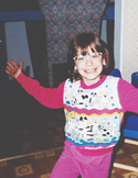 A smiling young girl with glasses and a colorful sweater spreads her arms wide in a happy pose.