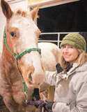 A smiling woman in a green hat standing next to a light-colored horse in a stable setting.