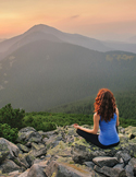 A woman with long red hair sits in a meditative pose on a rocky mountain peak overlooking a vast valley.