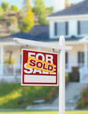 A red "For Sale" sign with a yellow "Sold" sticker in front of a blurred white suburban house.