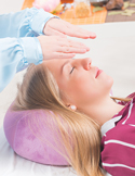 A Reiki practitioner holds their hands over the forehead of a woman lying down with her eyes closed in relaxation.