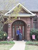 A man and woman standing together in front of a brick house with an arched wooden front door.