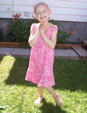 A smiling young girl without hair wearing a pink patterned dress stands happily in a sunny backyard.