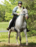 A woman wearing a black safety helmet smiling while riding a white horse through a grassy field.