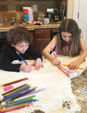 Two children sitting at a kitchen counter focused on drawing with colored pencils on large sheets of paper.
