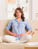 A woman sitting cross-legged on a sofa with her eyes closed, hands resting on her stomach in a meditative pose.