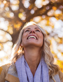 A smiling woman looks upward toward sunlight filtering through the golden leaves of a tree in autumn.