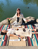 A woman in white leads a healing session by a lake for another person lying on a blanket.