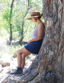 A woman in a sun hat leans peacefully against the thick, textured trunk of a large tree in a forest.