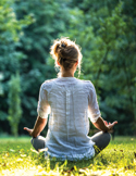 A woman sits in a meditative yoga pose in a sunlit, lush green forest clearing.