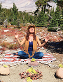 A woman meditating with raised hands in a mountain landscape behind a small rock altar.