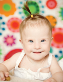 A smiling baby with Down syndrome and a small ponytail sitting in front of a colorful, floral background.