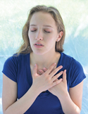 A woman in a blue shirt with eyes closed, holding her hands over her heart in a peaceful or emotional gesture.
