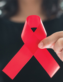 Close-up of a person's hand holding a red awareness ribbon against a dark, blurred background.