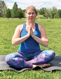 Woman sitting on a yoga mat in a grassy field, eyes closed with hands in a prayer position.