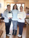 Three smiling women standing indoors, two of whom are holding official-looking certificates.