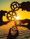 Multiple hands holding wooden gears against a sunset, symbolizing teamwork and alignment.