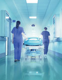 Two medical professionals push a gurney down a bright, sterile blue-toned hospital corridor.