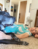A smiling woman lies on a carpeted floor next to a large, grey-muzzled black dog in a bright living room.