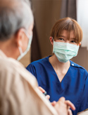 A medical professional in blue scrubs and a face mask looking kindly at an elderly patient.