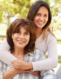 A smiling mother and daughter hugging outdoors, wearing light-colored sweaters in a sunlit park.