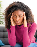 A woman sitting on a couch with her eyes closed, pressing her fingers to her temples to relieve a headache.
