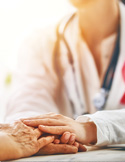 A doctor in a white coat compassionately holding an elderly patient’s hand in a brightly lit room.