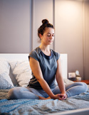 A woman sitting cross-legged on a bed in a meditative pose with her eyes closed.