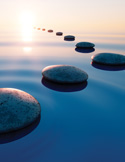 Stepping stones create a path across calm, blue water toward a glowing sunset.