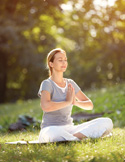 A woman sits in a meditative cross-legged pose on grass in a sunlit park with her hands in prayer.