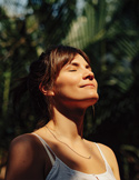 Close-up of a woman with her eyes closed, basking in warm sunlight with leafy shadows in the background.