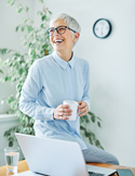 A joyful older woman with glasses laughs while sitting on a desk next to her laptop with a coffee mug.