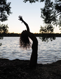 Silhouette of a person stretching backward by a peaceful lake at sunset, framed by dark tree branches.