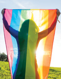 A person stands in a sunny field holding up a large, vibrant pride flag behind them.
