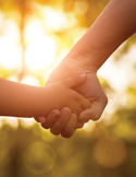Close-up of an adult and child holding hands against a warm, golden sunset background.