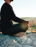 A man sits in a meditative pose on a rocky cliff during a golden sunset, focusing on mindfulness.