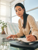 A woman sitting on a couch, focused on a laptop while writing notes in a black journal.