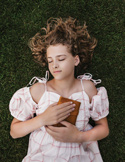 A girl lying on green grass with her eyes closed, holding a journal to her chest in a peaceful pose.