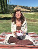 A woman sitting on a rug outdoors, holding a bowl and looking upward in a prayerful, peaceful pose.