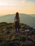 Woman standing on a rocky mountain ridge at sunset, looking out over layered distant hills and a golden sky.
